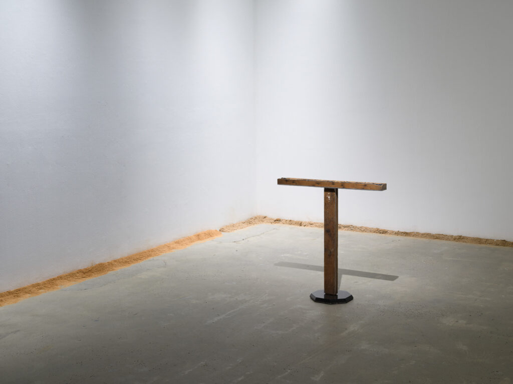 This is a color photograph of two artworks by Lukas Geronimas. In the foreground to the right and featured at a slight angled view is a sculpture entitled Custom T, composed of two rectangular pieces of wood with one mounted perpendicular on top of the other to form the shape of a T. The wooden T is mounted to a black octagonal plexiglass base. In the background and along the perimeter of the floor and walls is another artwork titled Puzzledust, which is sawdust that was produced, collected and stored by the artist over the past two years.