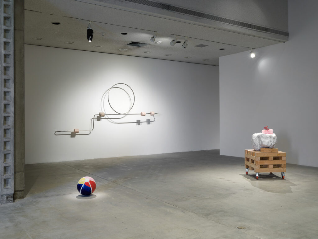 This is a color photograph of three sculptures by Nevine Mahmoud. Pictured left to right are: a sculpture titled Beach Ball IV, which is a multi-colored spherical ceramic work placed on the floor and resembles an inflatable beach ball; a wall-mounted sculpture titled Abacus arm 1, which features a linear steel bar with a circle and four stone components. Each of these components are wrapped around the steel bar and are different stones, such as onyx, Portuguese marble and Aphrodite marble; and a sculpture titled Three Components, which includes a large white boulder with a small, pink, bulbous glass object that has a red coil placed on top of it. These particular elements sit on top of a large stack of wood pieces on wheels.