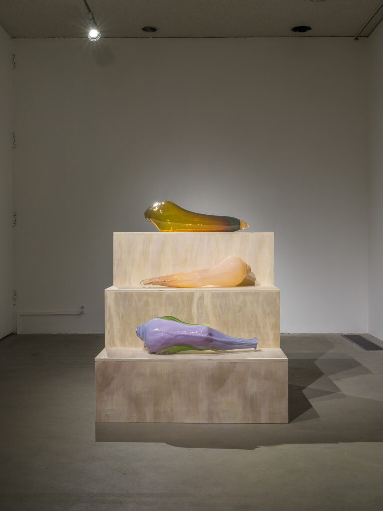 This is a color photograph of three large blown glass sculptures of roughly equal size by Nevine Mahmoud resting on a tiered, wooden three step pedestal. At the top is a sculpture entitled yellow Toy, an amber colored blown glass piece resembling a human leg. On the center tier is a sculpture entitled Leg (Lila), a flesh toned blown glass piece resembling a human leg. Beneath this on the last tier is another leg shaped work called Leg (lilac), made of lilac and green colored blown glass.