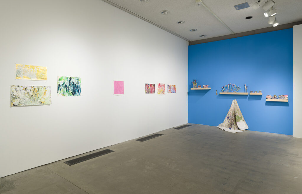 This is a color photograph of two walls in an exhibition of Vanessa McConnell's artworks at the Los Angeles Municipal Art Gallery. On the right white wall are seven brightly colored abstract paintings on paper by Vanessa McConnell. On the far right is a grouping of three, two featuring the use of yellow and green paint mounted one on top of the other on the left, and on the right a third painting featuring mostly blue and green paint. On the center of the left wall is a painting featuring primarily pink paint. On the right side of this wall is another grouping of three paintings, featuring mostly shades of red, yellow, and blue paint. On the right blue wall are four floating shelves containing a variety of paint-covered objects, including paintbrushes and various cups and dishes. In the center of the wall underneath one of the shelves is a paint splattered fabric drop cloth, draped on a single hook flowing down the wall and resting on the floor.
