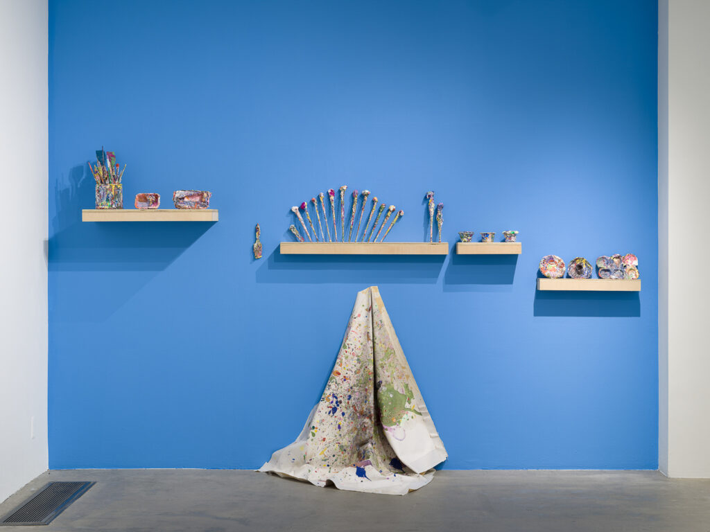 This is a color photograph of a blue wall in an exhibition space of Vanessa McConnell's artworks at the Los Angeles Municipal Art Gallery. There are four floating shelves mounted to the wall, and on each shelf lies paint-covered ephemera, including paint brushes, cups, dishes, and mixing trays. In the center of the wall under one of the shelves is a light colored paint slpattered drop cloth, hung from a single hook draped against the wall and resting on the floor.