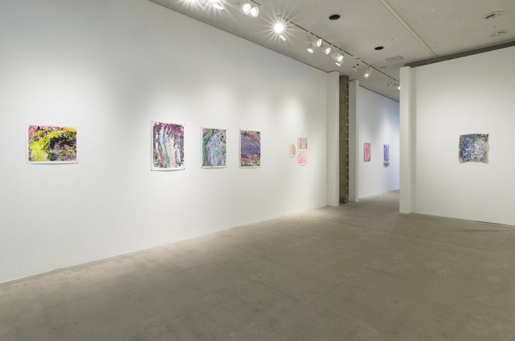 This is a color photograph of part of an exhibition of Vanessa McConnell's artworks at the Los Angeles Municipal Art Gallery. On the left wall are seven brightly colored abstract paintings on paper by Vanessa McConnell. On the far left is a painting featuring pink, yellow, and black paint in rough, dynamic brushstrokes. In the center of the wall are three paintings of equal size, consisting of various shades of blue, pink, purple, and green. On the right side of the wall are three small paintings grouped closely together in lighter shades of paint featuring mostly vertical brushstrokes. On the right wall is a single abstract painting on a piece of burlap featuring shades of blue, white, and green. Through an opening in this wall, two abstract paintings of roughly equal size can be seen on a wall in the distant background.